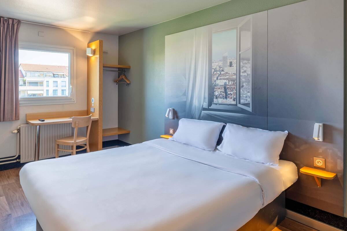 Quarto de hotel com uma cama grande, uma escrivaninha, uma cadeira e vista para o horizonte urbano através de uma janela.
