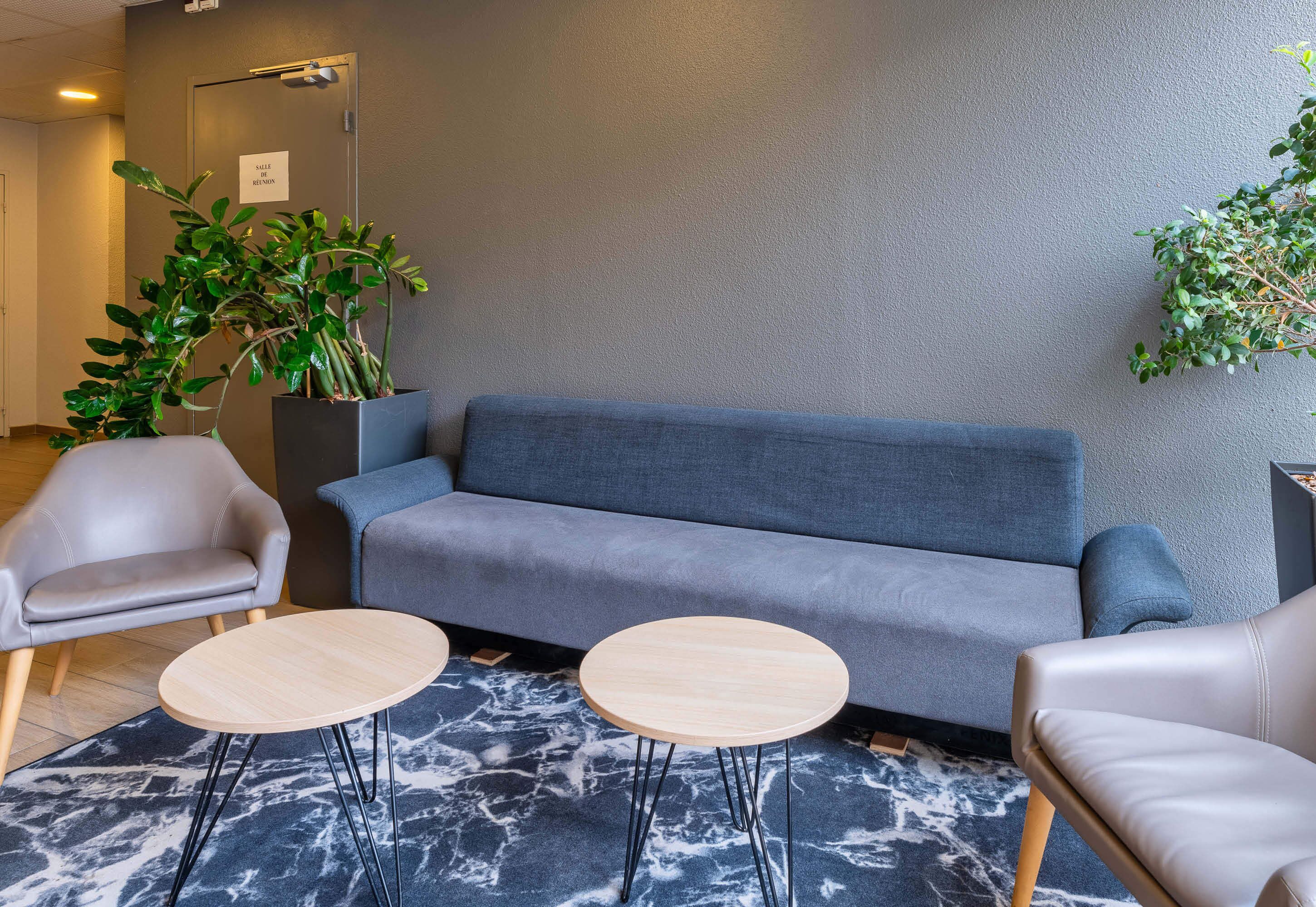 Un área de descanso moderna con un sofá gris, dos mesas redondas de madera, un sillón gris y una planta en maceta.