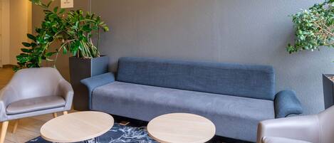 Un área de descanso moderna con un sofá gris, dos mesas redondas de madera, un sillón gris y una planta en maceta.