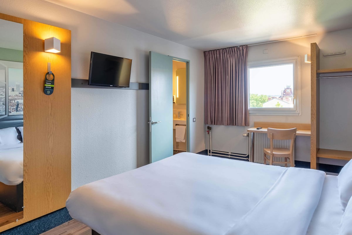 Quarto de hotel com cama, escrivaninha, cadeira, televisão e janela com cortinas.