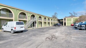 A parking lot with a white van and several cars parked in front of a building with arched windows and a staircase leading to an upper level. - Sienna Inn Channelview, TX (Channelview)