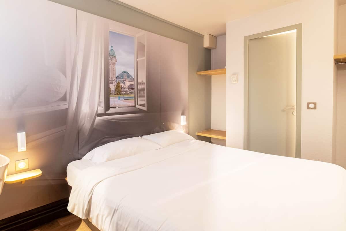 A hotel room with a bed, a window with curtains, a nightstand with a lamp, and a door.