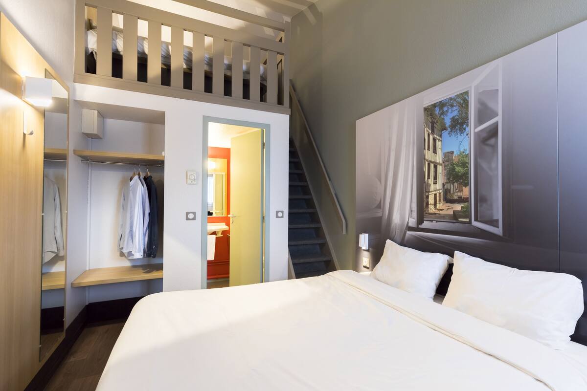 A hotel room with a bed, a wardrobe, a mirror, a window with a view, and a staircase.