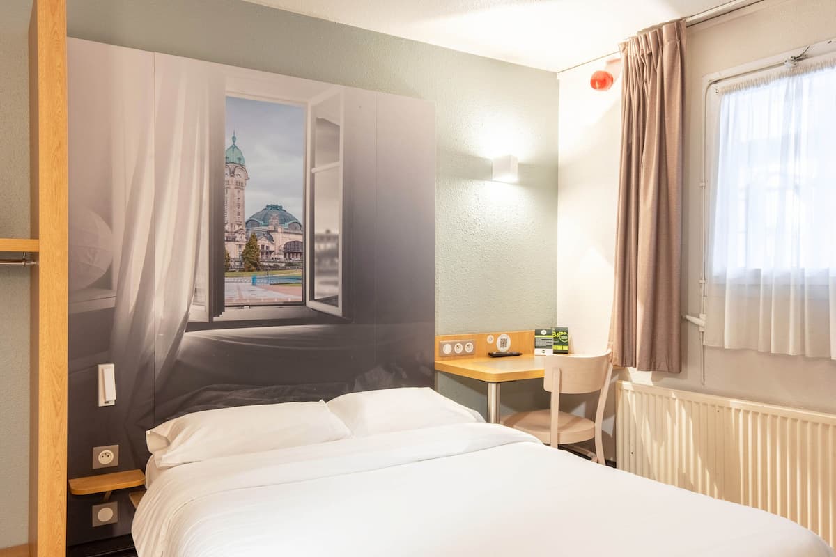 A hotel room with a bed, a desk, a chair, a radiator, and a window with curtains.