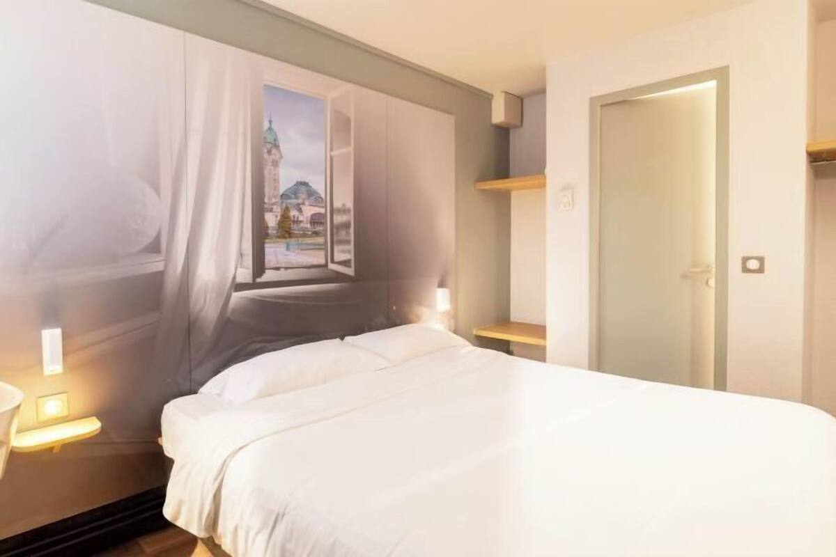 A hotel room with a bed, a window with curtains, a door, and a nightstand with a lamp.