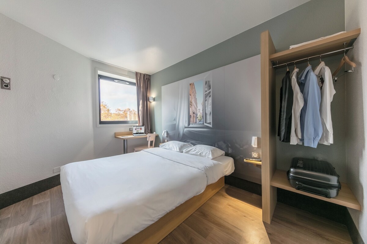 A modern hotel room with a single bed, a desk, a chair, a wardrobe, and a window with curtains.
