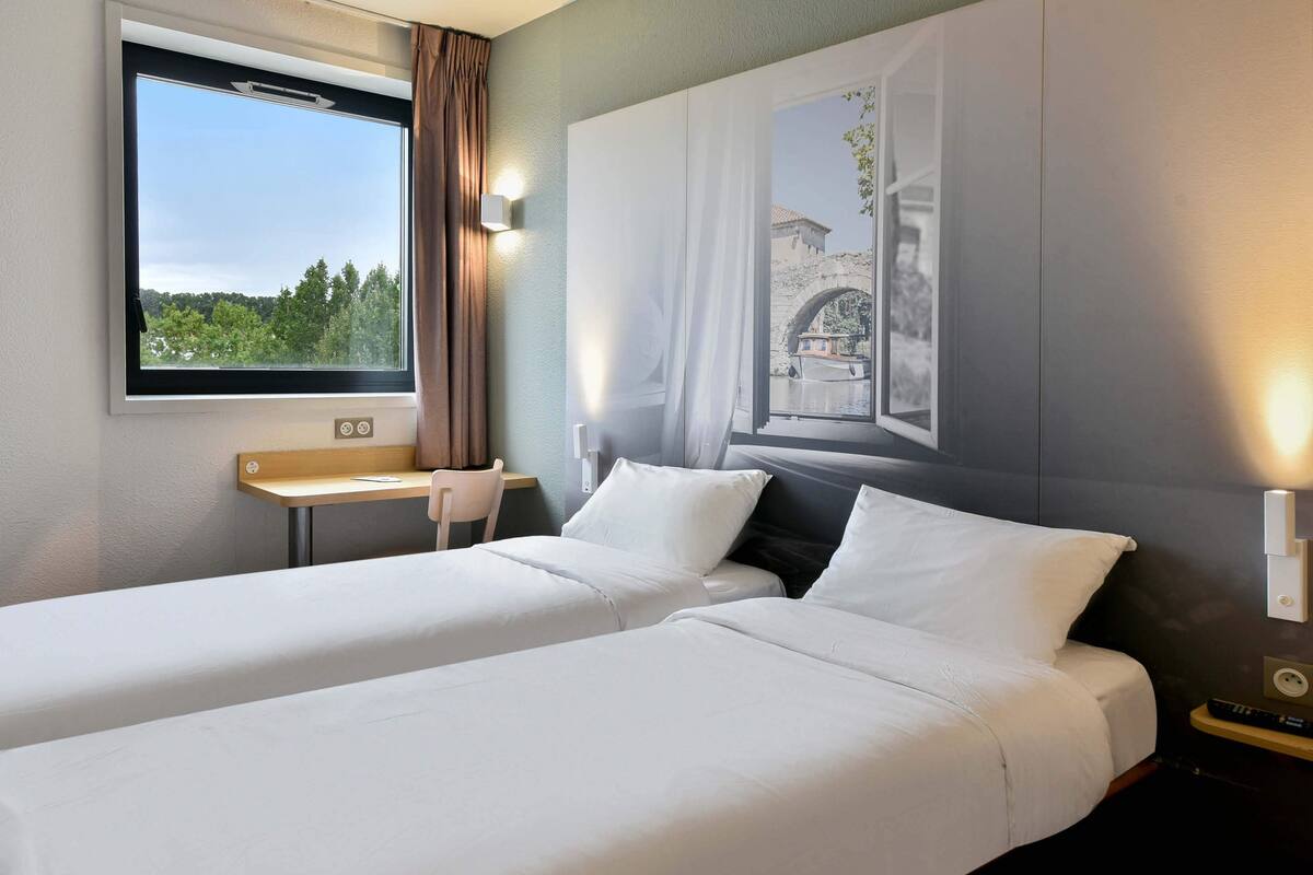 A hotel room with two beds, a desk, and a window with a view of greenery and a building.