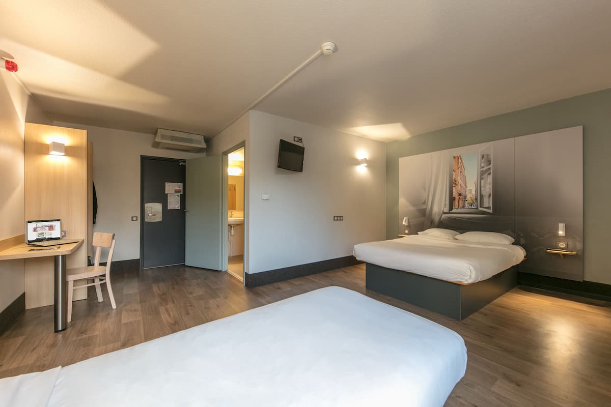 A hotel room with a bed, a desk, and a chair. There is a painting on the wall and a TV mounted on the wall.