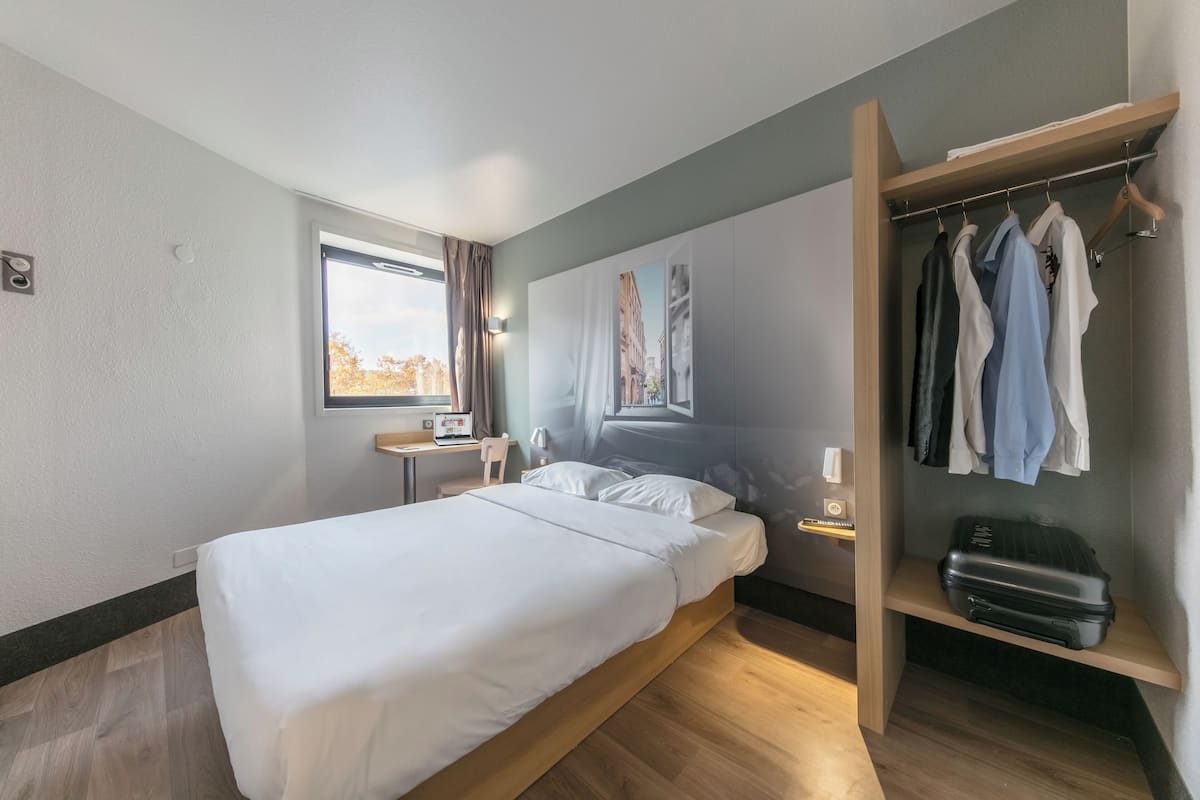 A hotel room with a bed, a desk, a chair, a wardrobe, and a window with curtains.