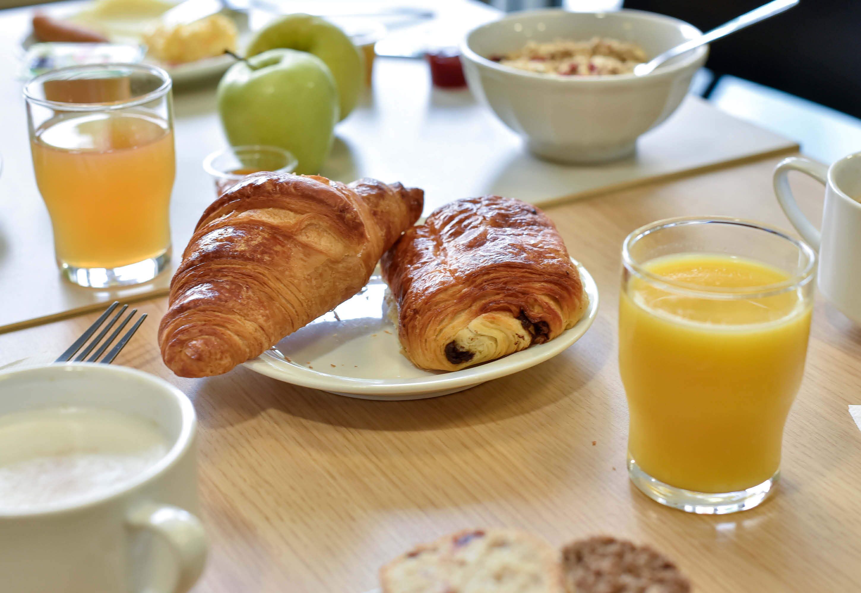 Una mesa de desayuno con croissants, un vaso de zumo de naranja, un tazón de cereal y una manzana verde.