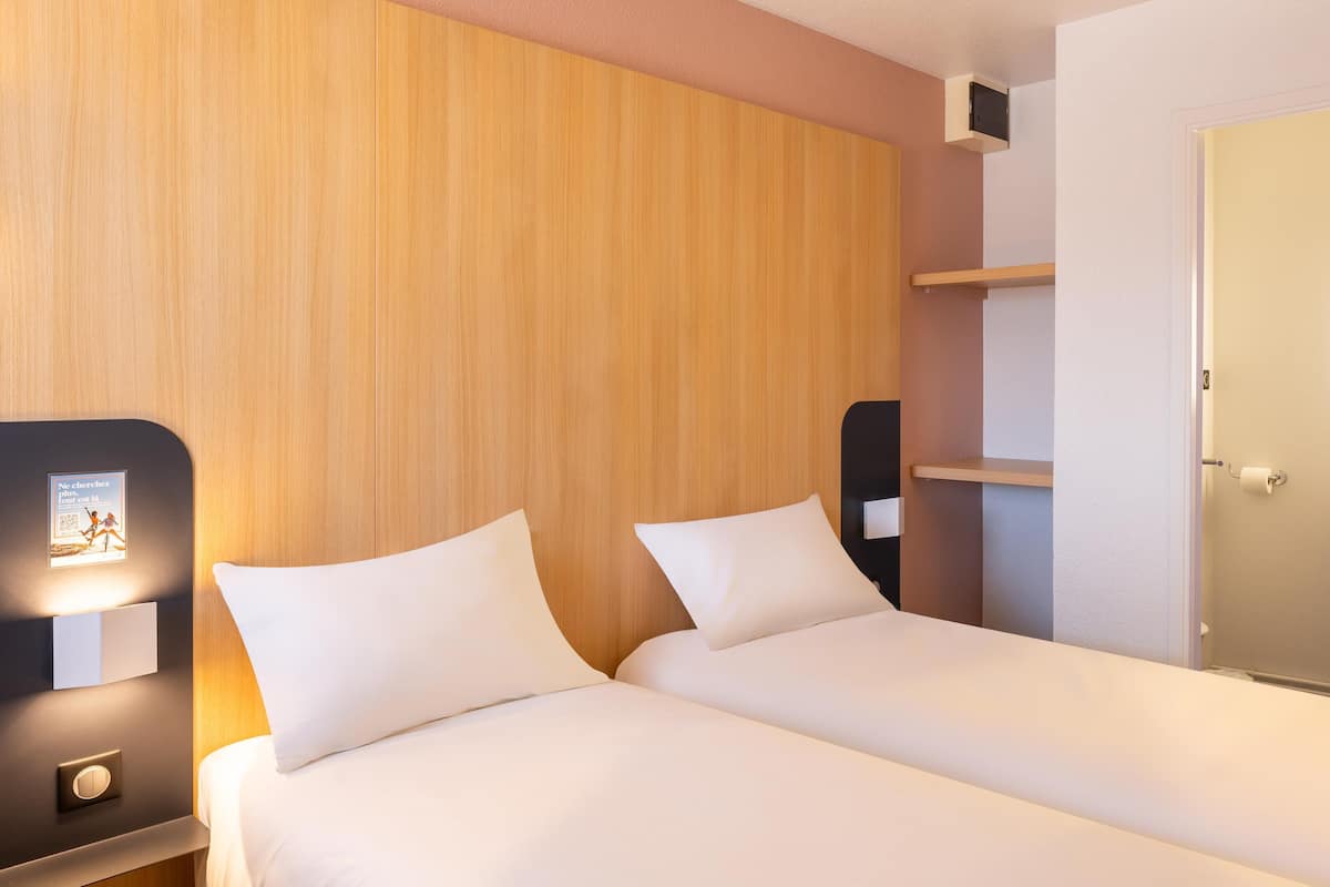 A hotel room with two beds, a wooden headboard, a bedside table with a lamp, a small shelf, and a door leading to a bathroom.