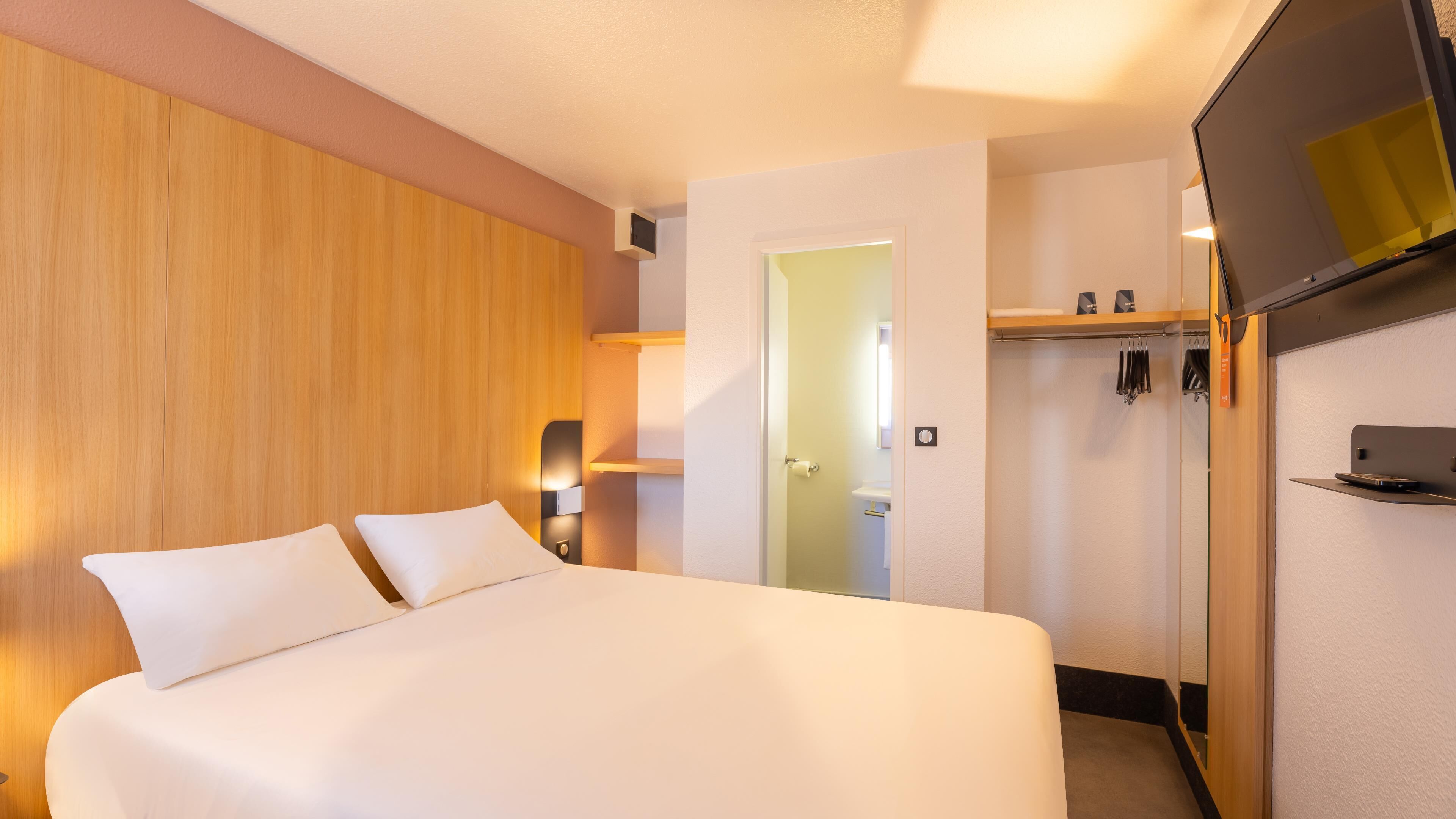 A hotel room with a bed, a wooden panel wall, a door, a coat rack, and a small shelf.