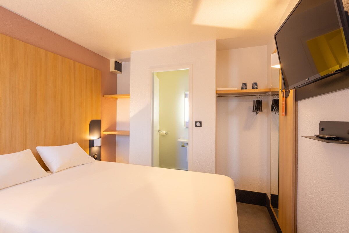 A hotel room with a bed, a wooden panel wall, a door, a coat rack, and a small shelf.