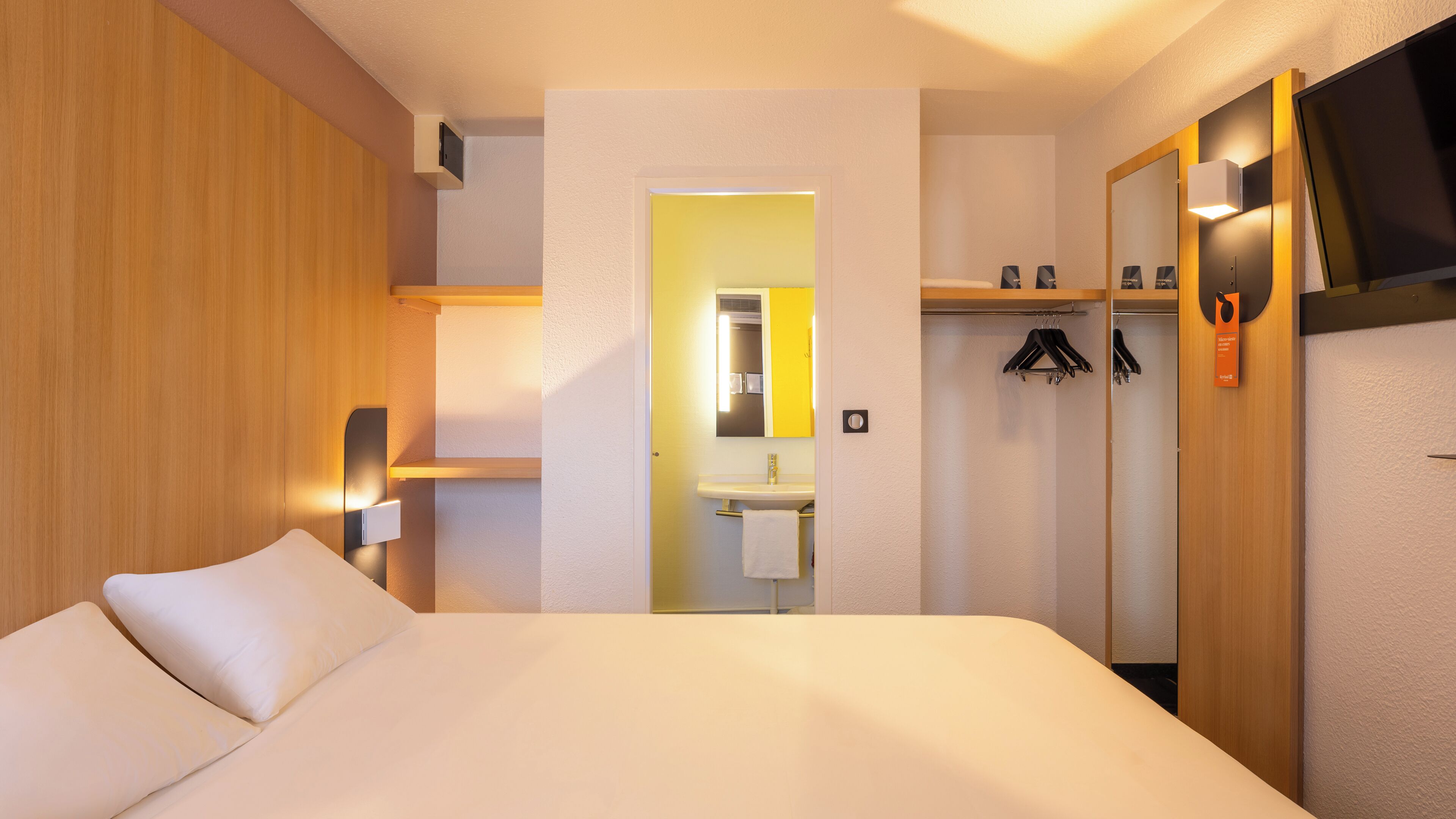 Habitación de hotel con cama, mesita de noche, espejo, armario y baño visible a través de una puerta abierta.