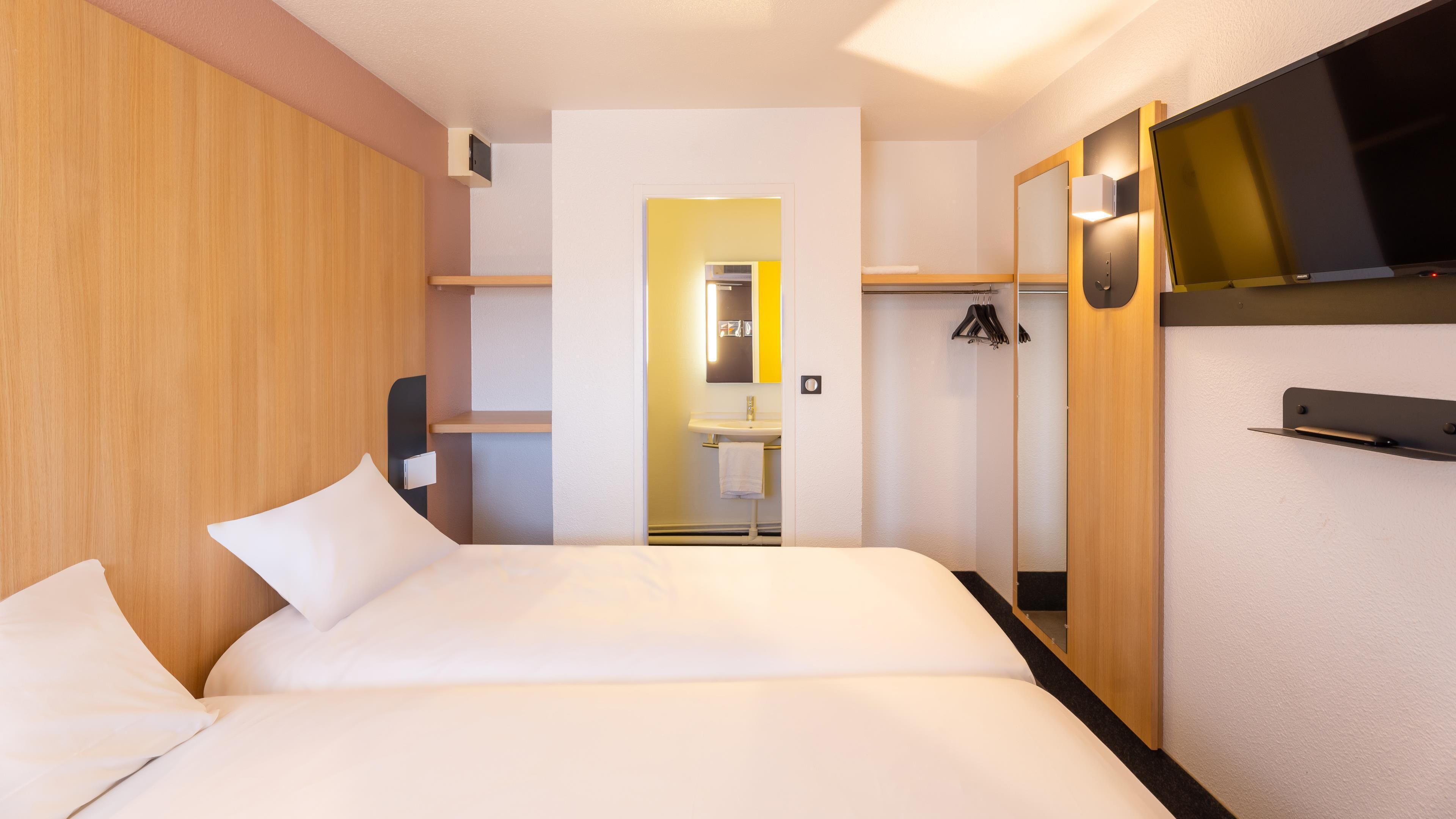 A hotel room with a bed, a mirror, a TV, and a small bathroom with a sink and towel rack.
