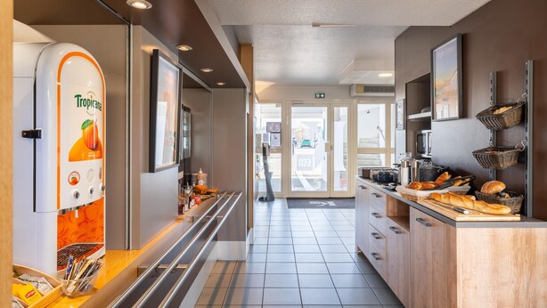 A modern hotel kitchen with a self-service beverage station, a buffet counter with bread and pastries, and a view of the street outside. - Kyriad ECO - Lorient Lanester (Lanester)