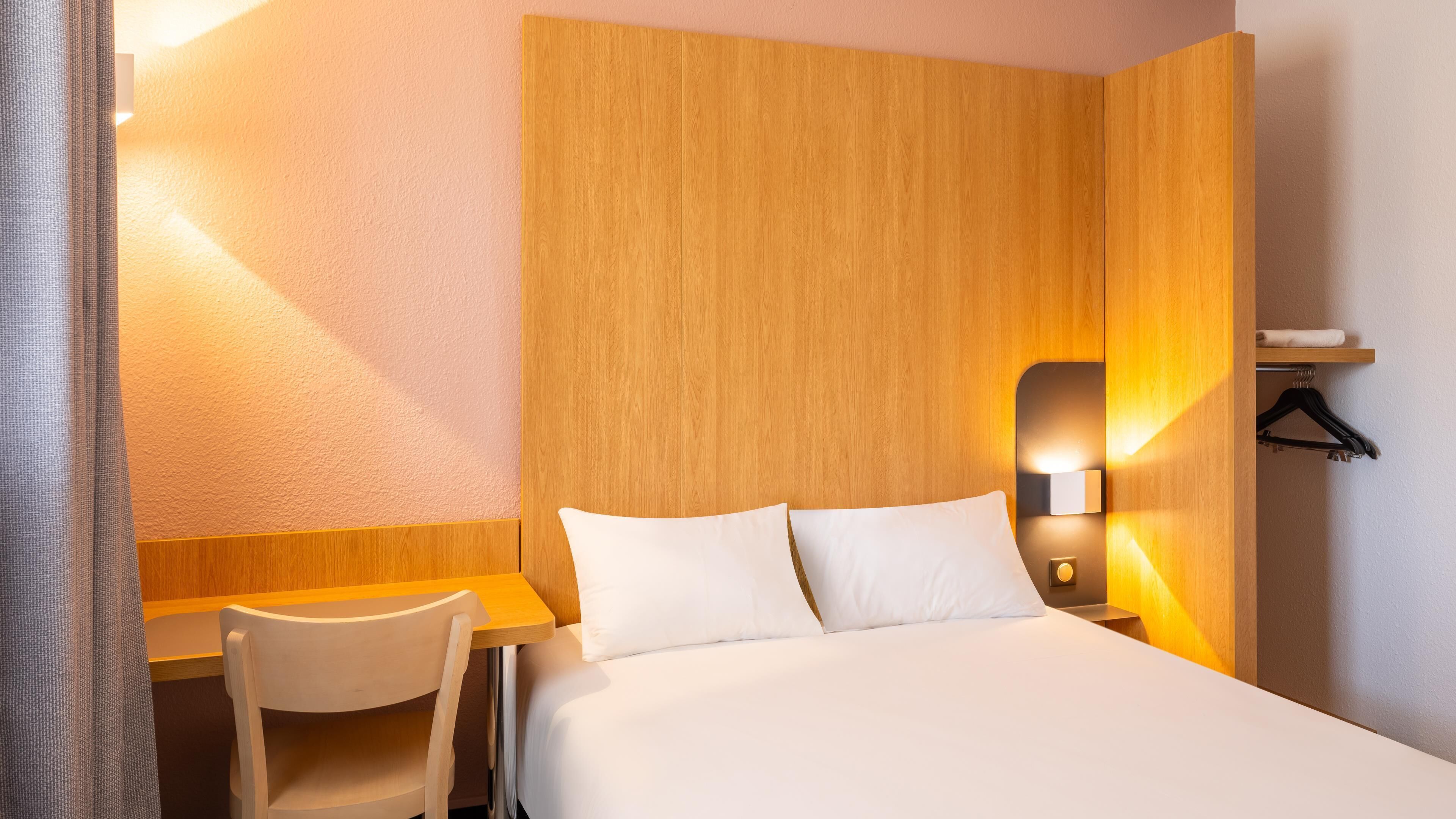 A hotel room with a bed, a desk, a chair, a wooden headboard, a wall-mounted light, and a clothing hanger.