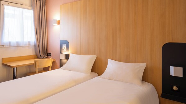 A hotel room with two beds, a desk, a chair, a wooden headboard, and a wall-mounted light.
