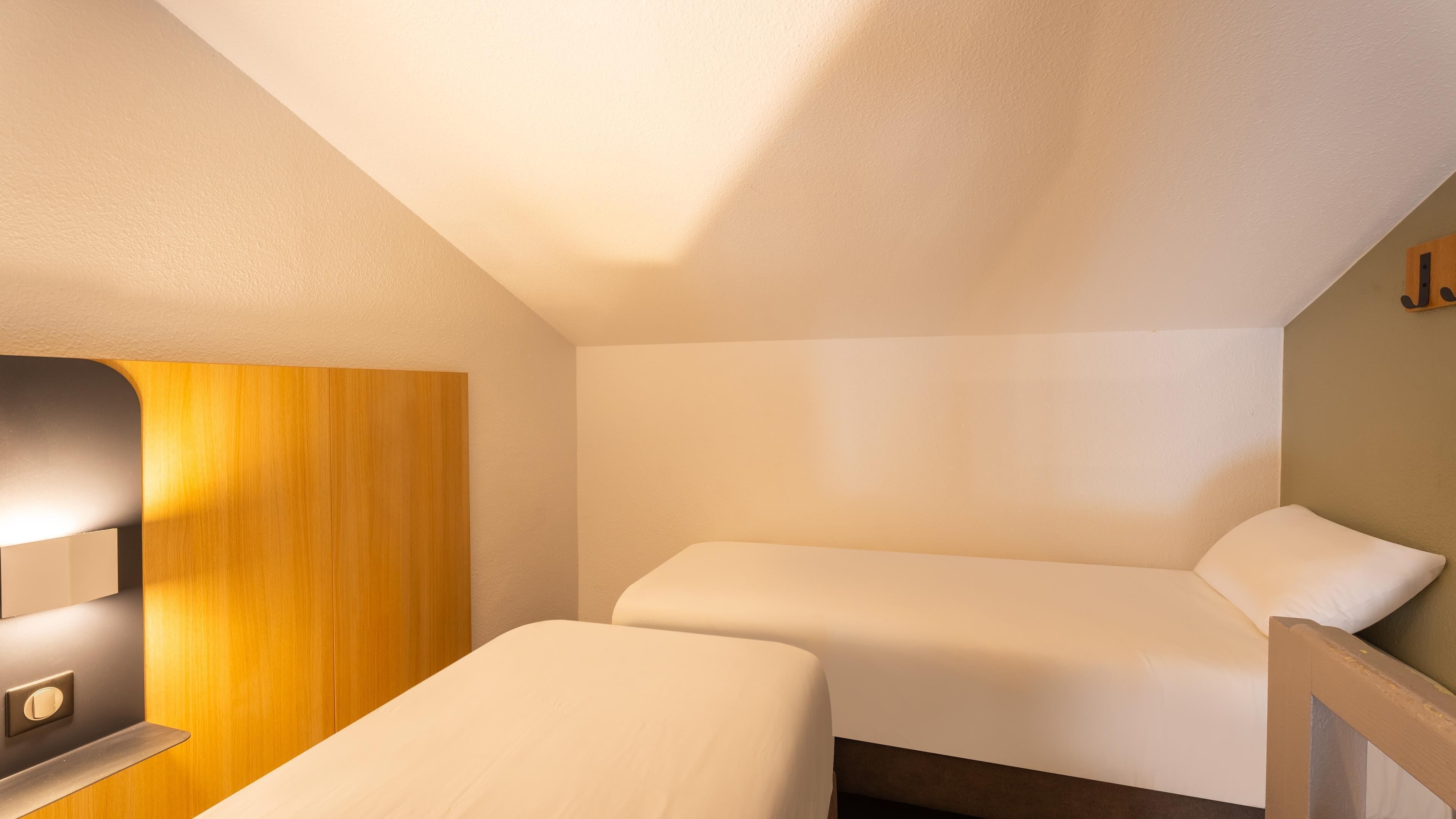 Une petite pièce bien éclairée, avec deux lits, un mur recouvert de panneaux de bois et une petite table avec une lampe.