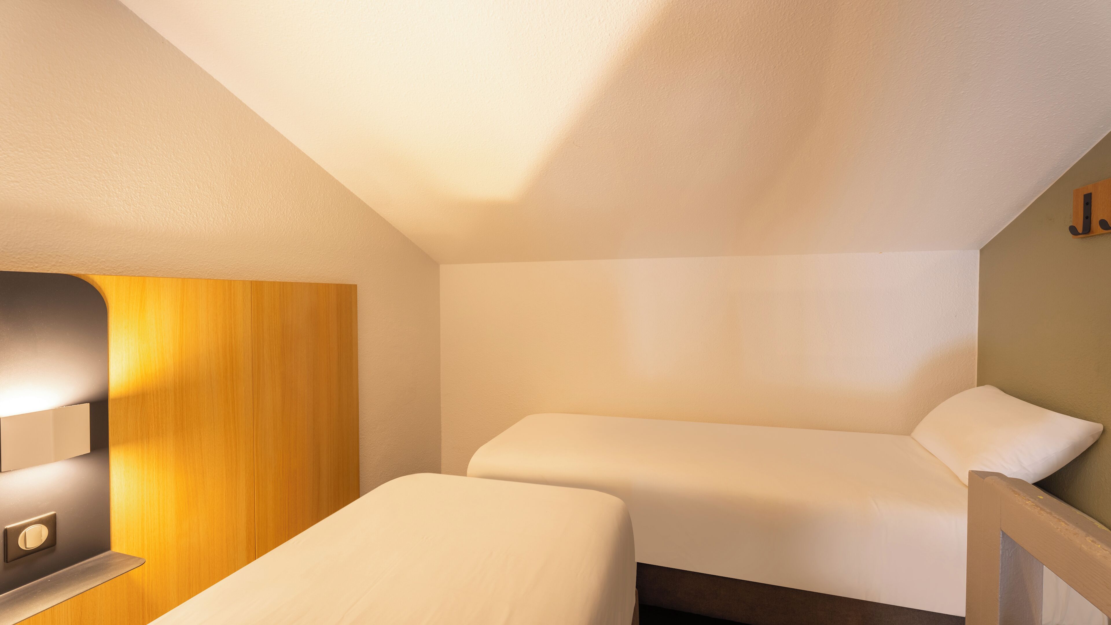 Una habitación pequeña y luminosa con dos camas, un panel de madera en la pared y una mesita de noche.