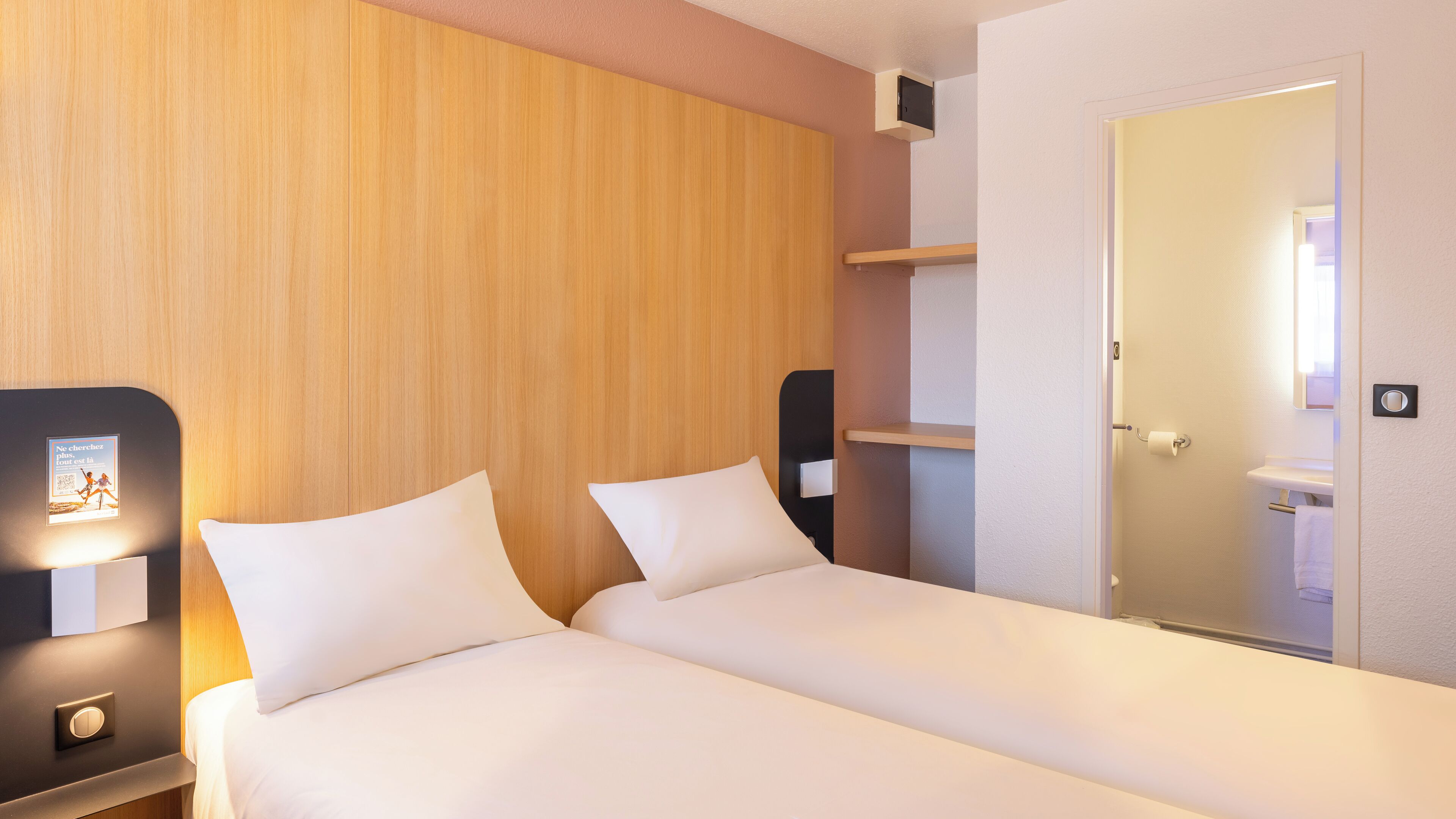 Une chambre d’hôtel avec une tête de lit en bois, deux lits avec du linge de lit blanc, une table de chevet avec une lampe, une étagère et une porte donnant sur une salle de bain.
