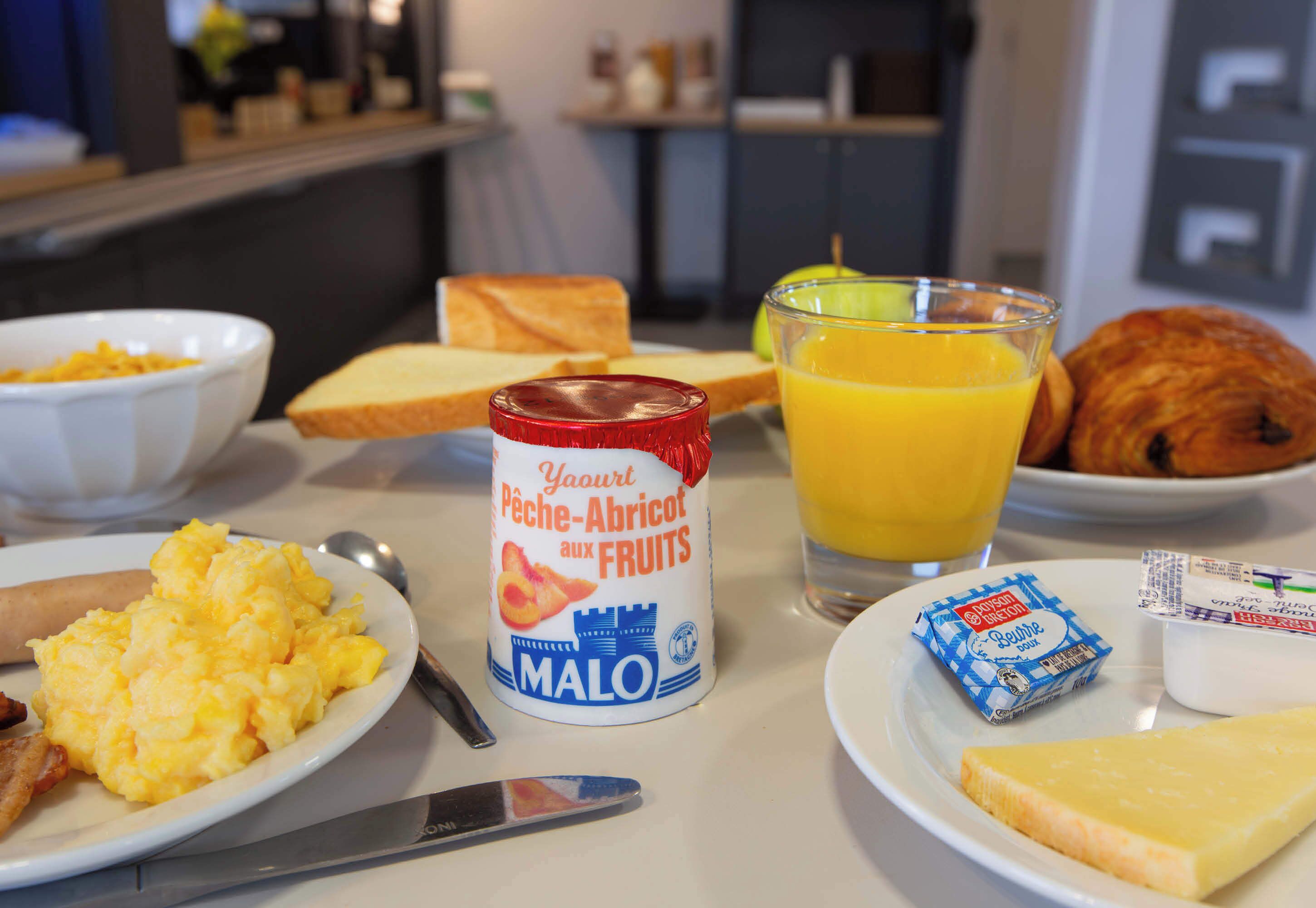 Una mesa de desayuno con huevos revueltos, tostadas, un yogur de frutas en lata, un vaso de jugo de naranja, un croissant y una loncha de queso.