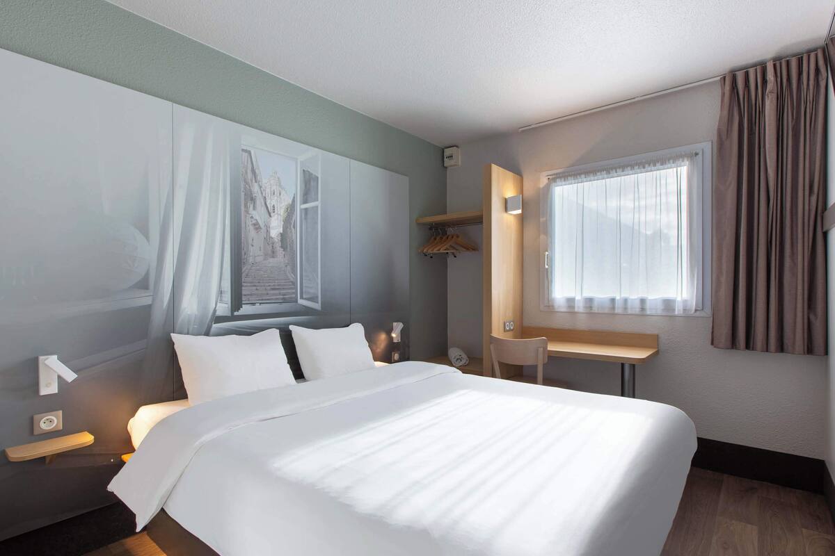 Ein Hotelzimmer mit einem großen Bett, einem Schreibtisch, einem Stuhl, einem Kleiderschrank und einem Fenster mit Vorhängen.