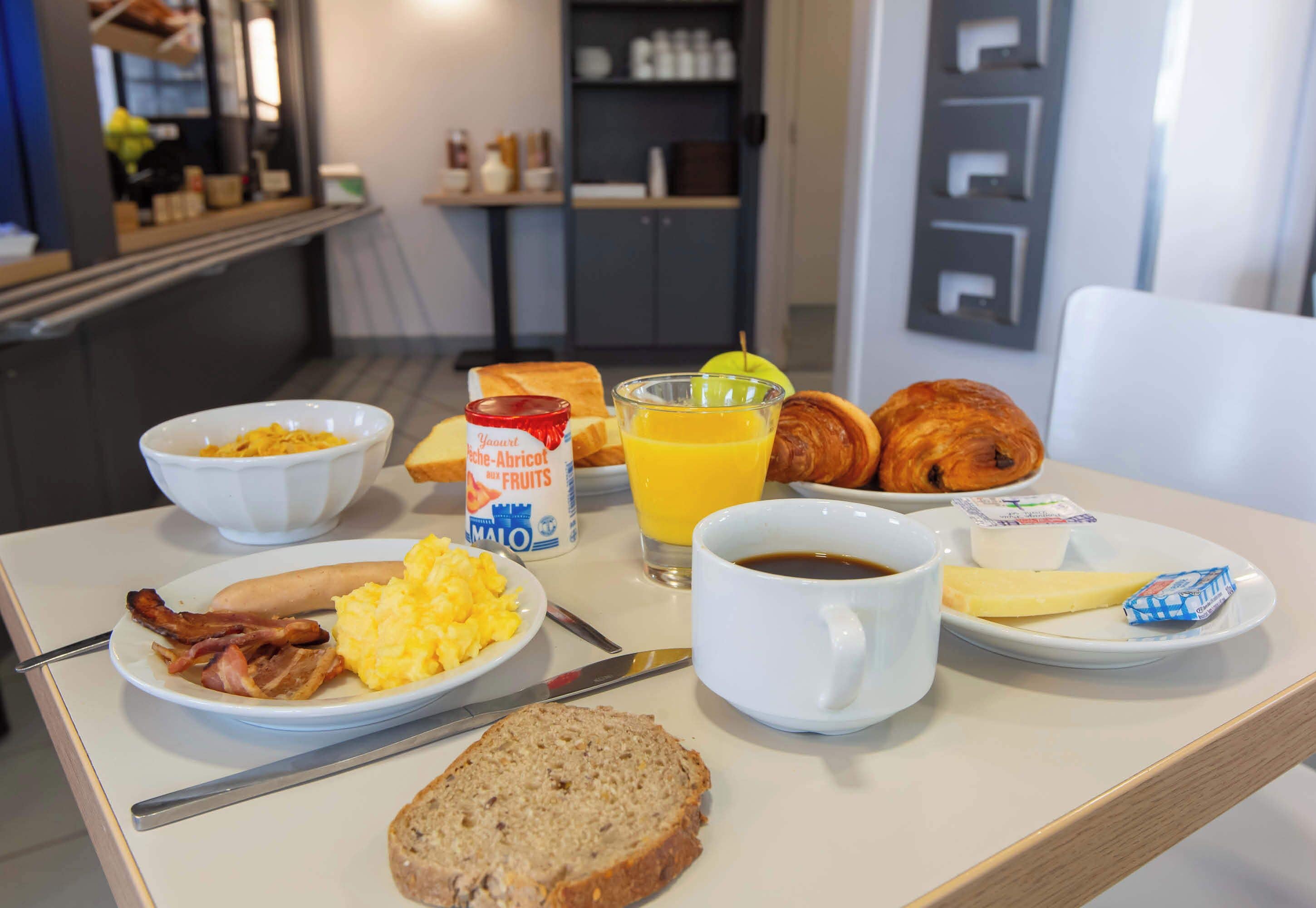 A breakfast table with scrambled eggs, bacon, toast, croissants, orange juice, coffee, and a bowl of cereal.
