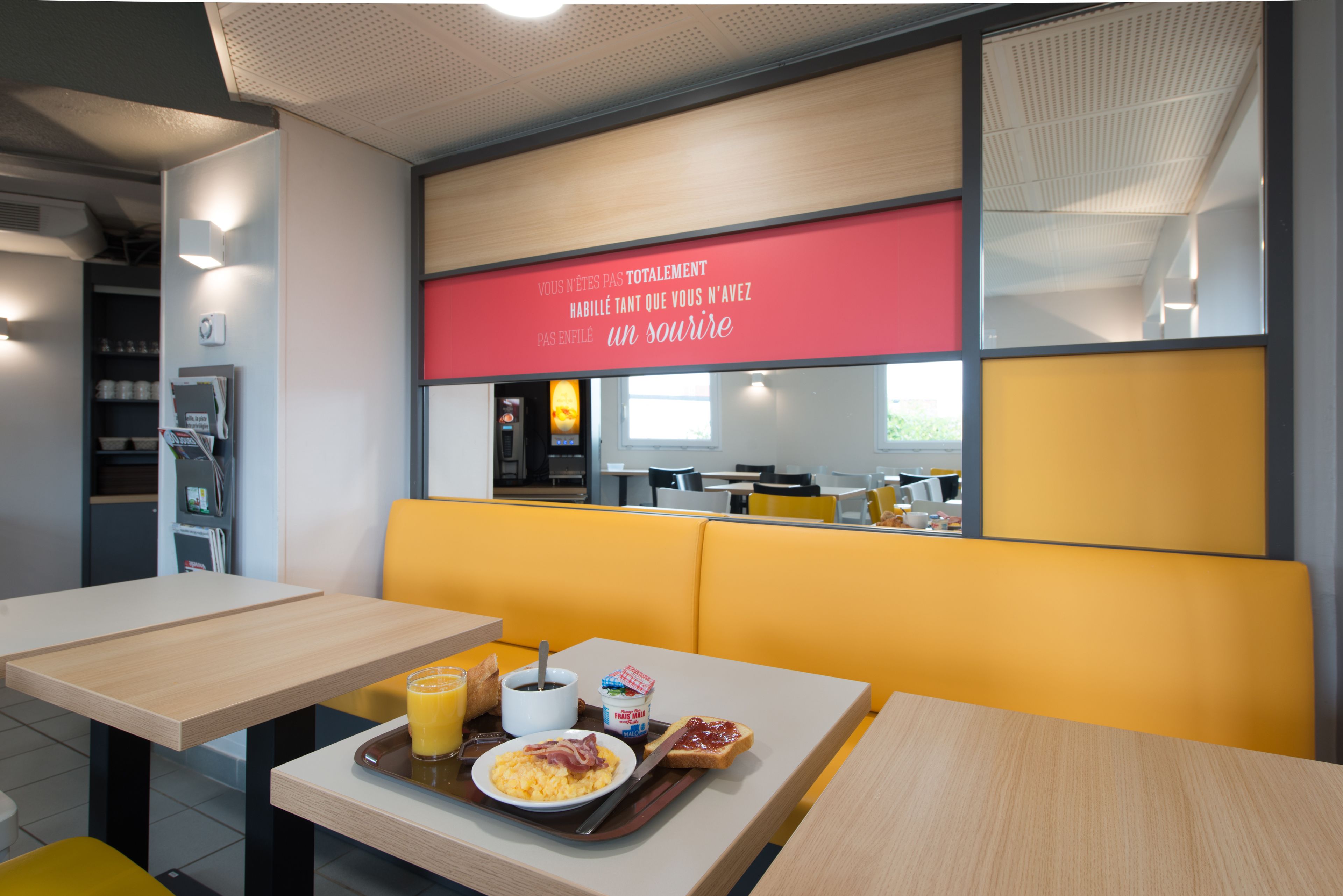 Una zona de dinar moderna amb seients tipus barra grocs, una taula de fusta amb articles per esmorzar, i un rètol vermell amb text en francès.