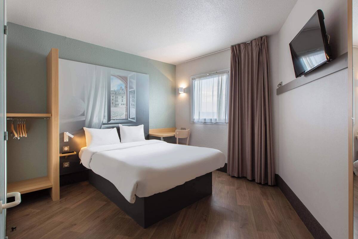 Ein Hotelzimmer mit einem großen Bett, Nachttischen, einem Schreibtisch, einem Sessel und einem Wandfernseher, sowie einem Fenster mit Vorhängen.