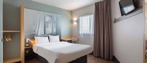 A hotel room with a large bed, bedside tables, a desk, a chair, a TV mounted on the wall, and a window with curtains.