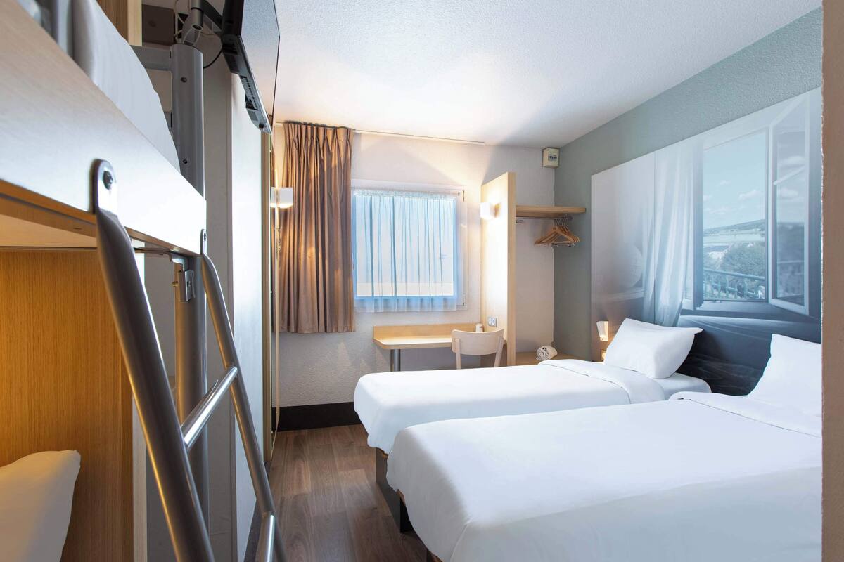 Ein Hotelzimmer mit zwei Betten, einem Schreibtisch, einem Stuhl, einem Kleiderhaken und einem Fenster mit Vorhängen.
