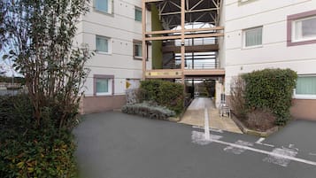 A building with a sign reading "ACCUEIL" indicating a reception area. There are multiple windows, a covered walkway, and a parking area with marked spaces.