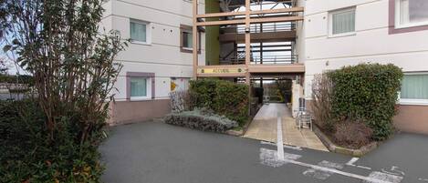 Un edificio con un cartel que dice “ACCUEIL”, indicando una zona de recepción. Se observan varias ventanas, una galería techada y un estacionamiento con espacios delimitados.