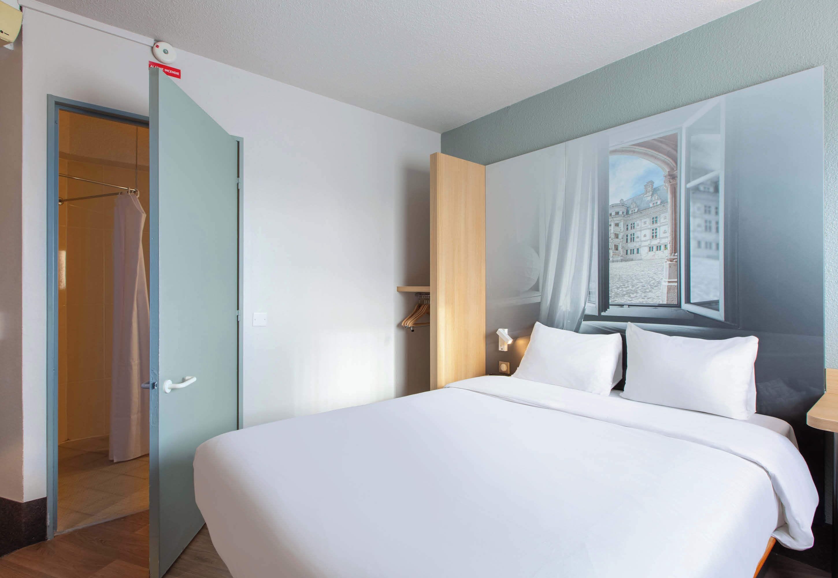 Habitación de hotel con una cama grande, un armario, un espejo y una ventana con vistas a edificios.