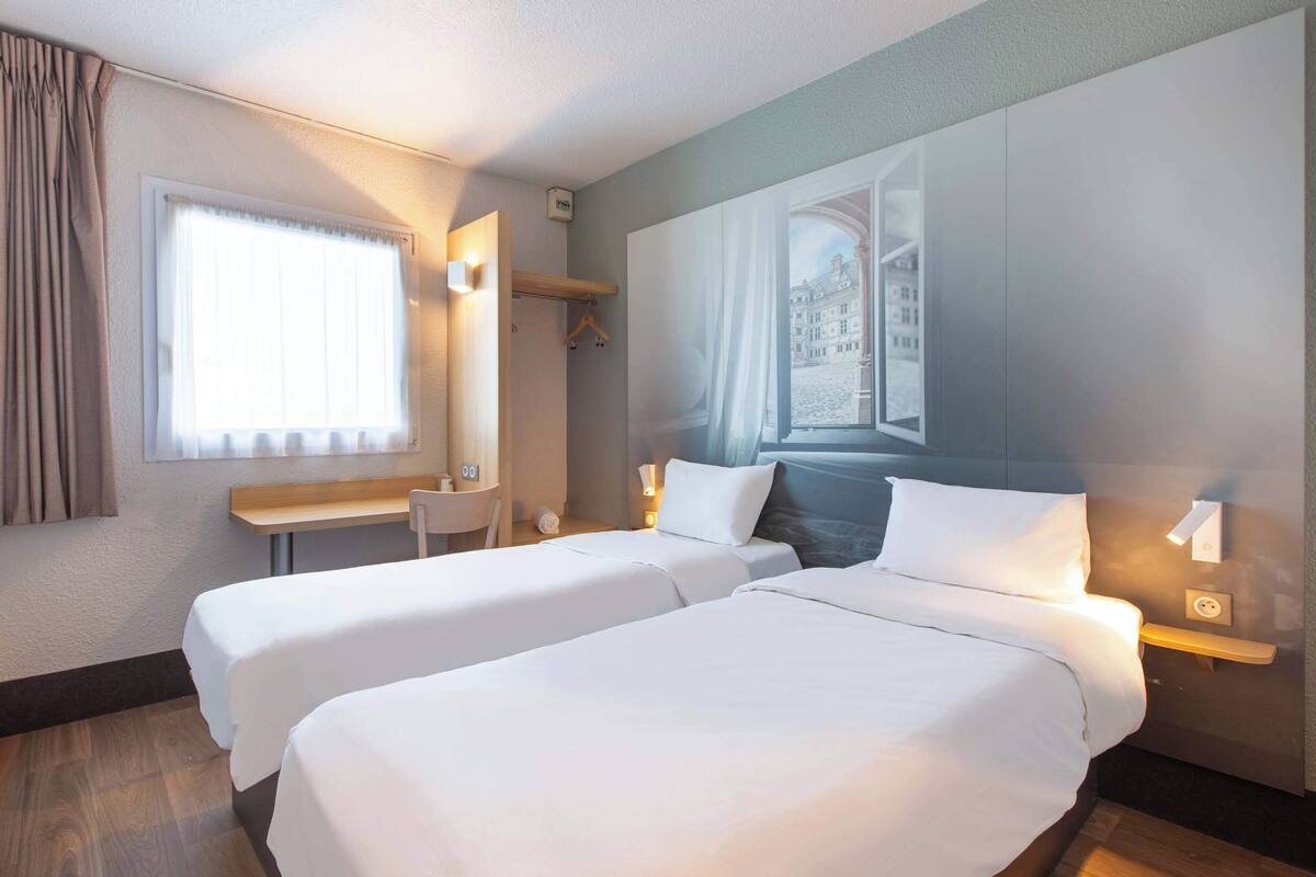 Ein Hotelzimmer mit zwei Betten, einem Schreibtisch, einem Stuhl und einem fenster mit Vorhängen.