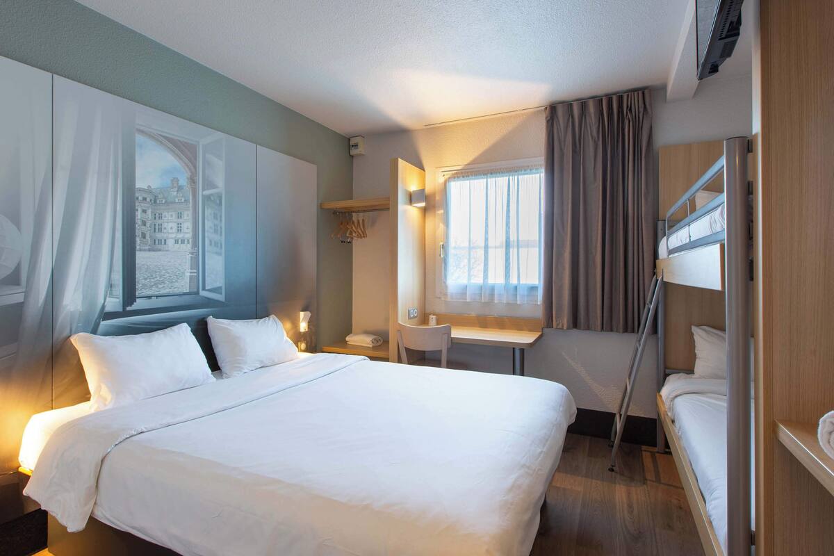 Ein Hotelzimmer mit einem großen Bett, einem Schreibtisch, einem Stuhl, einem Etagenbett und einem Fenster mit Vorhängen.