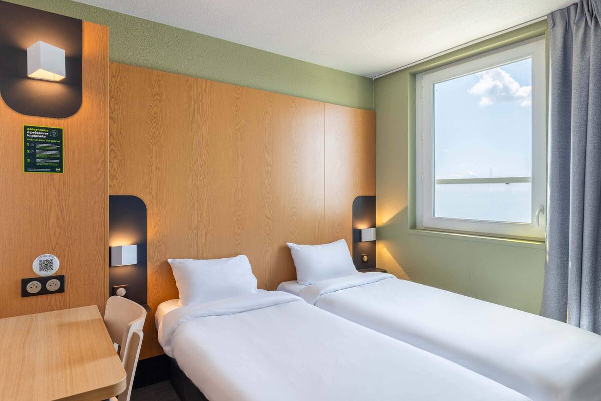 A hotel room with two beds, a wooden headboard, a small table, and a window with a view of the sea.