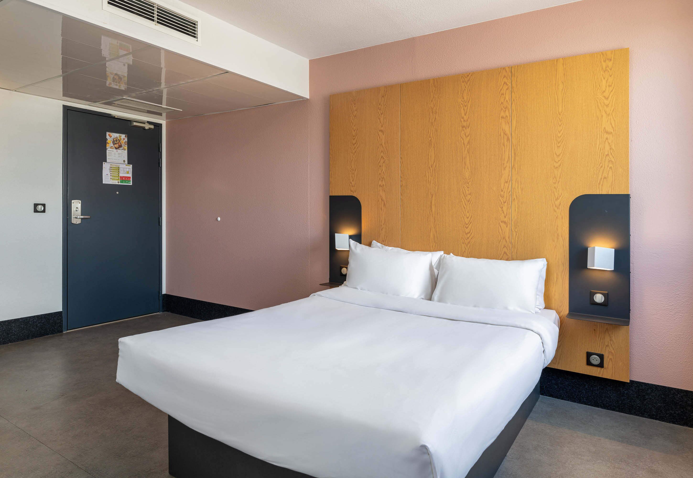 Une chambre d’hôtel avec un lit, une tête de lit en bois, deux lampes fixées au mur, une porte et une grille de ventilation fixée au mur.