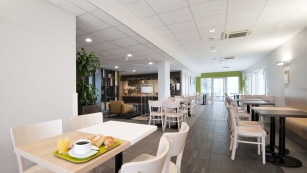 Un área de comedor moderna con mesas y sillas blancas, una estación de café y vista a la cocina al fondo.
