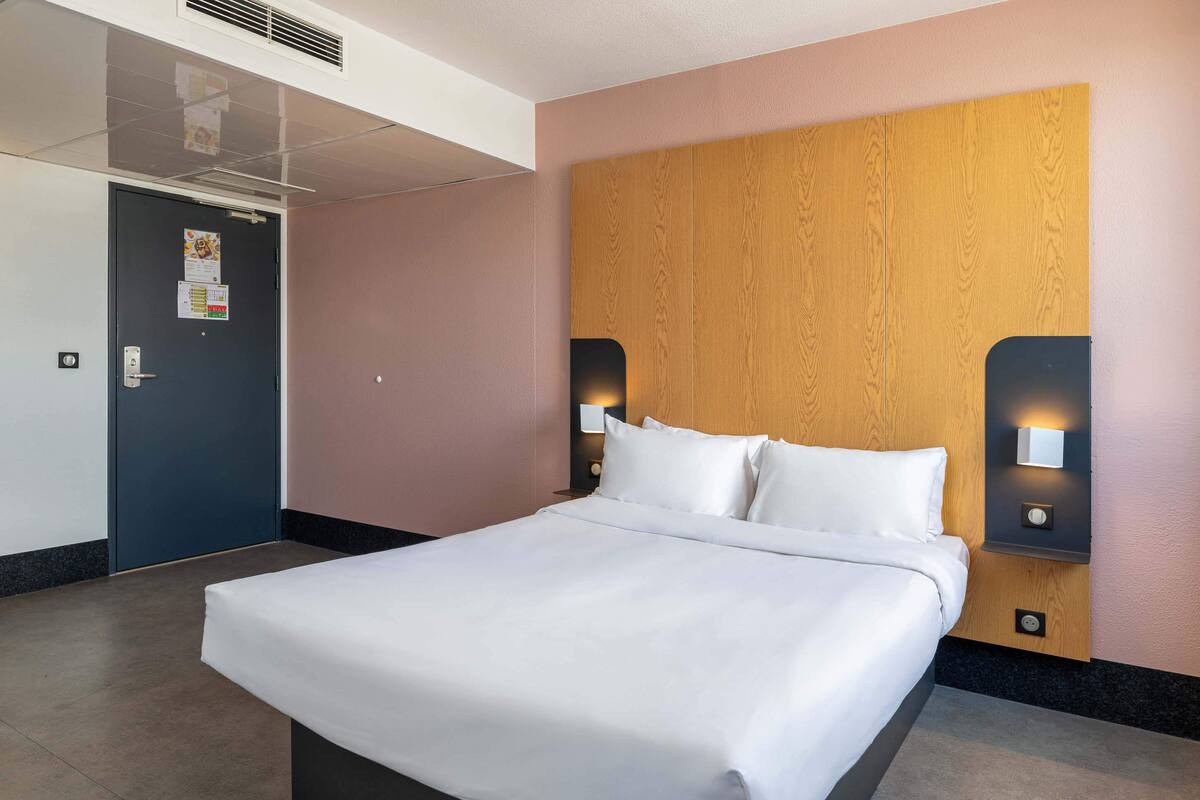 A hotel room with a large bed, wooden headboard, two wall-mounted lights, and a door with a calendar.