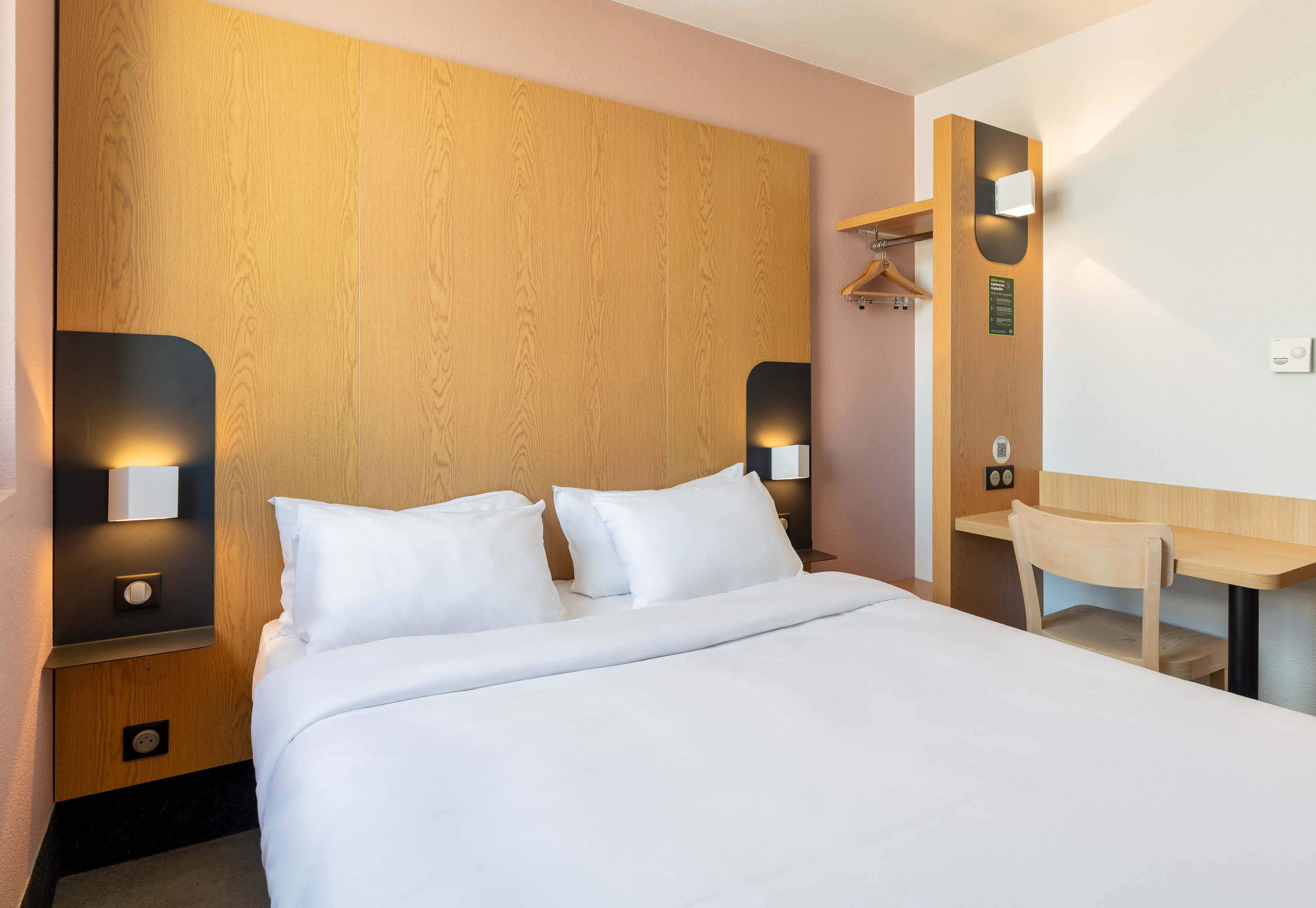 A modern hotel room with a wooden headboard, a bed with white linens, bedside tables with lamps, a wooden desk, and a mirror.