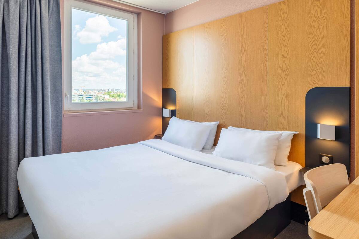 A hotel room with a large bed, a wooden headboard, two bedside lamps, a window with curtains, and a view of the cityscape.