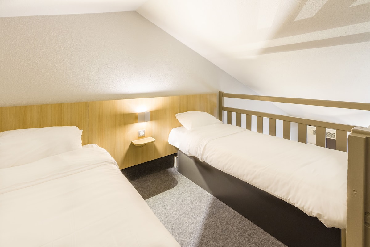 A small, well-lit room with two single beds, a wooden headboard, a bedside table with a lamp, and a carpeted floor.