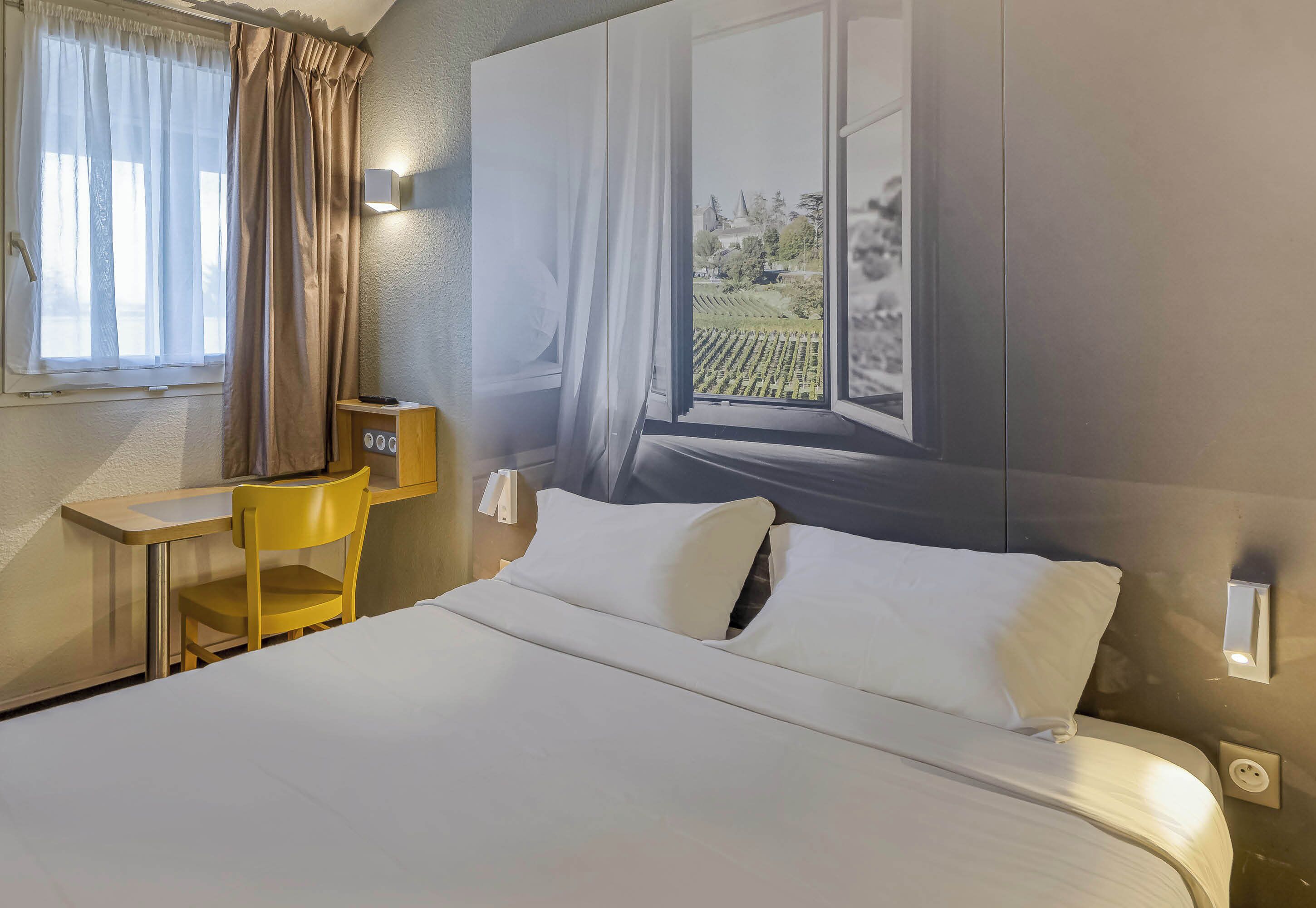 Ein Hotelzimmer mit einem Bett, einem Schreibtisch, einem Stuhl und einem fensterförmigen Ausschnitt mit Vorhängen.