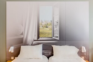 Un dormitorio con un ventanal grande, cortinas blancas, una cama con almohadas blancas y una mesita de noche con una lámpara.