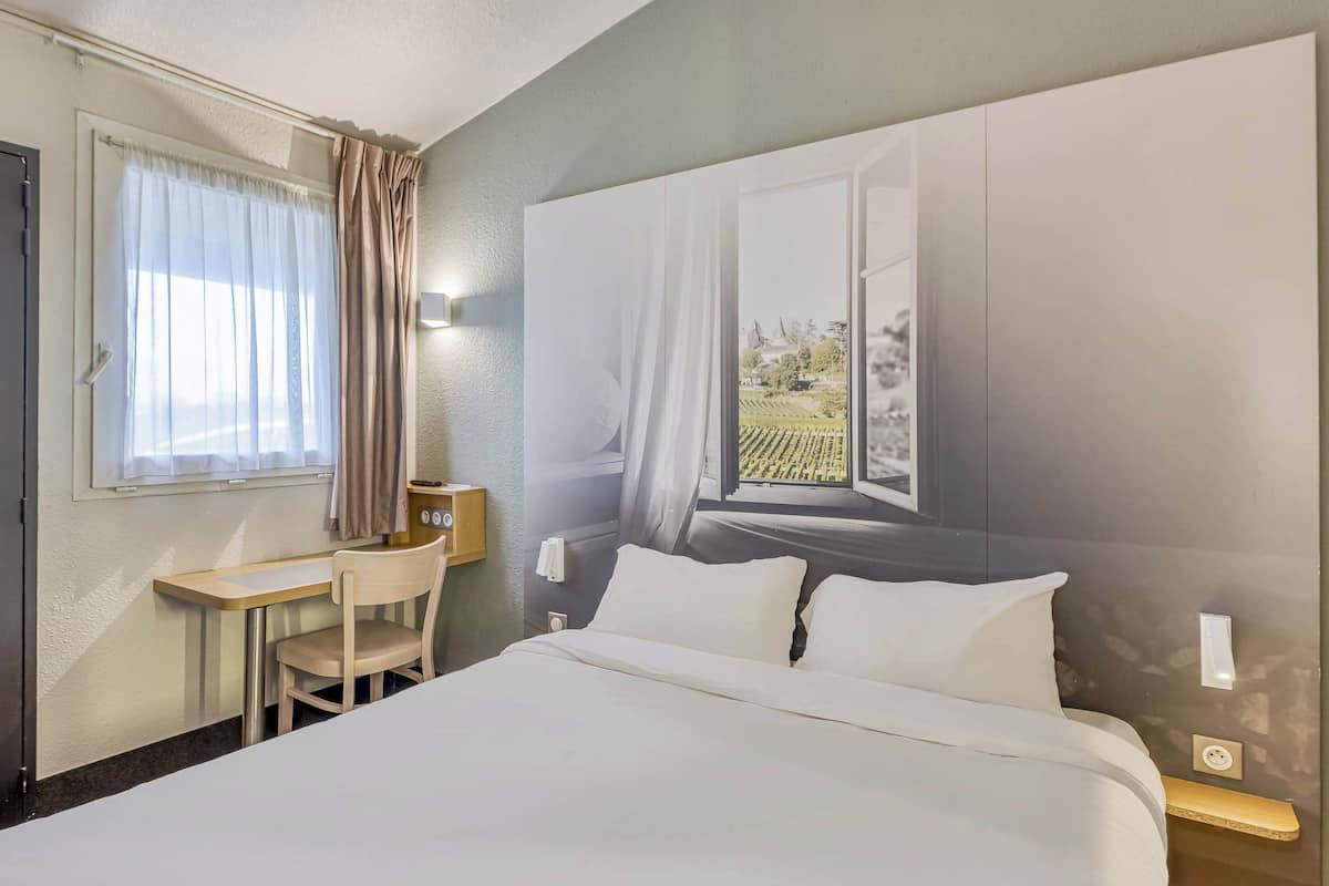 A hotel room with a bed, a desk, a chair, and a large window with curtains.