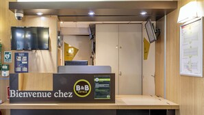 Una recepció amb un cartell que diu 'Bienvenue chez B&B Hotels', una càmera de seguretat, una televisió i un mapa a la paret.