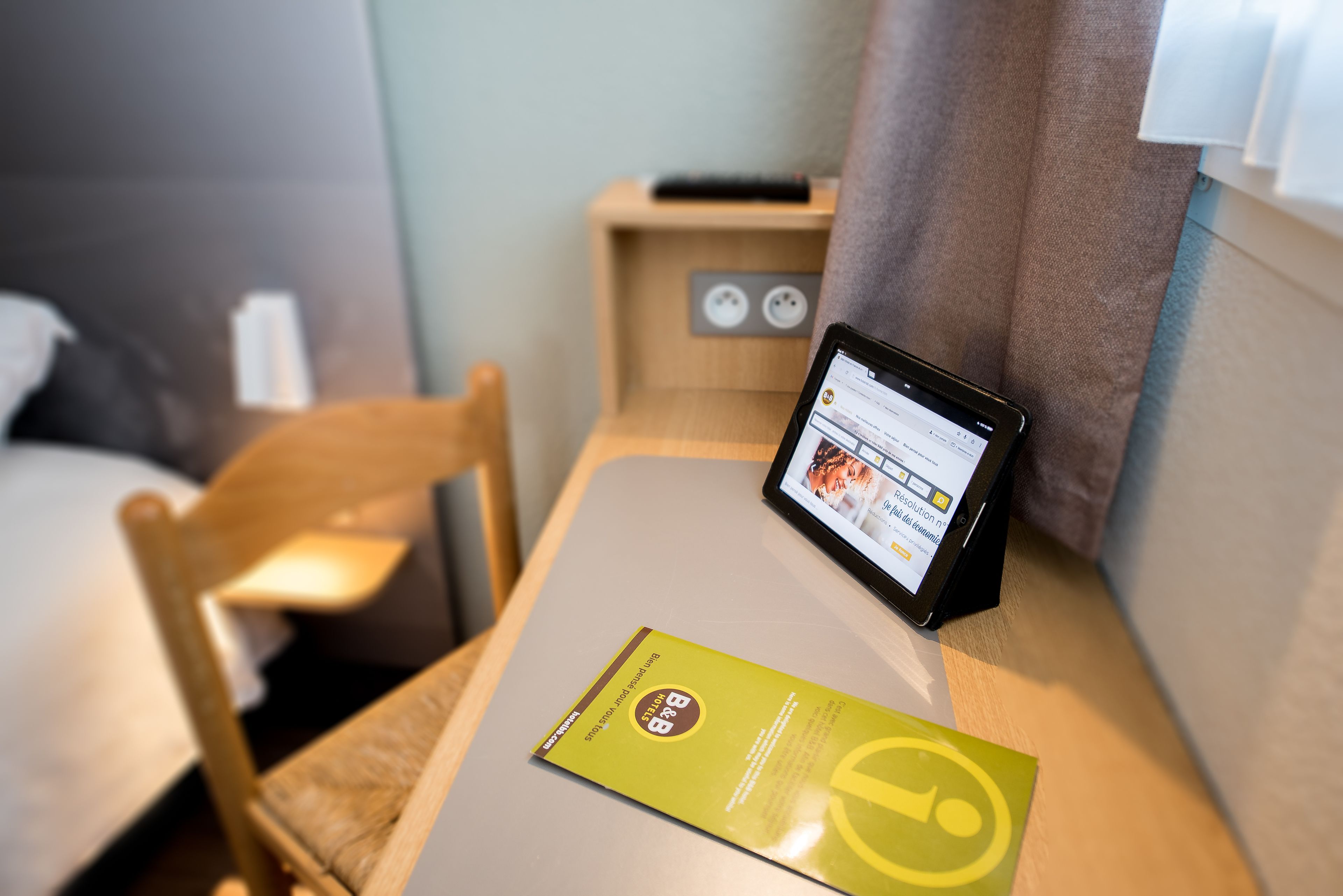 A tablet on a bedside table displaying a hotel app, a bed, a chair, and a wooden nightstand with a lamp.
