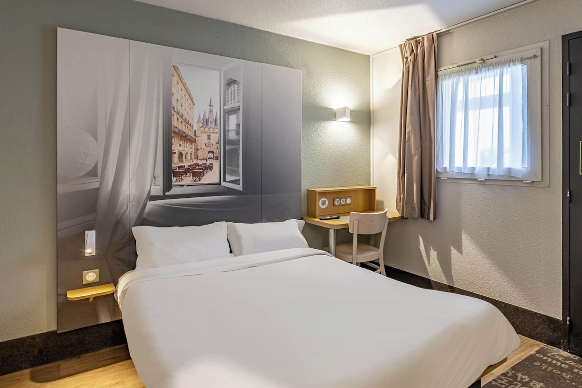 A hotel room with a large bed, a desk with a chair, a wall-mounted TV, and a window with curtains.